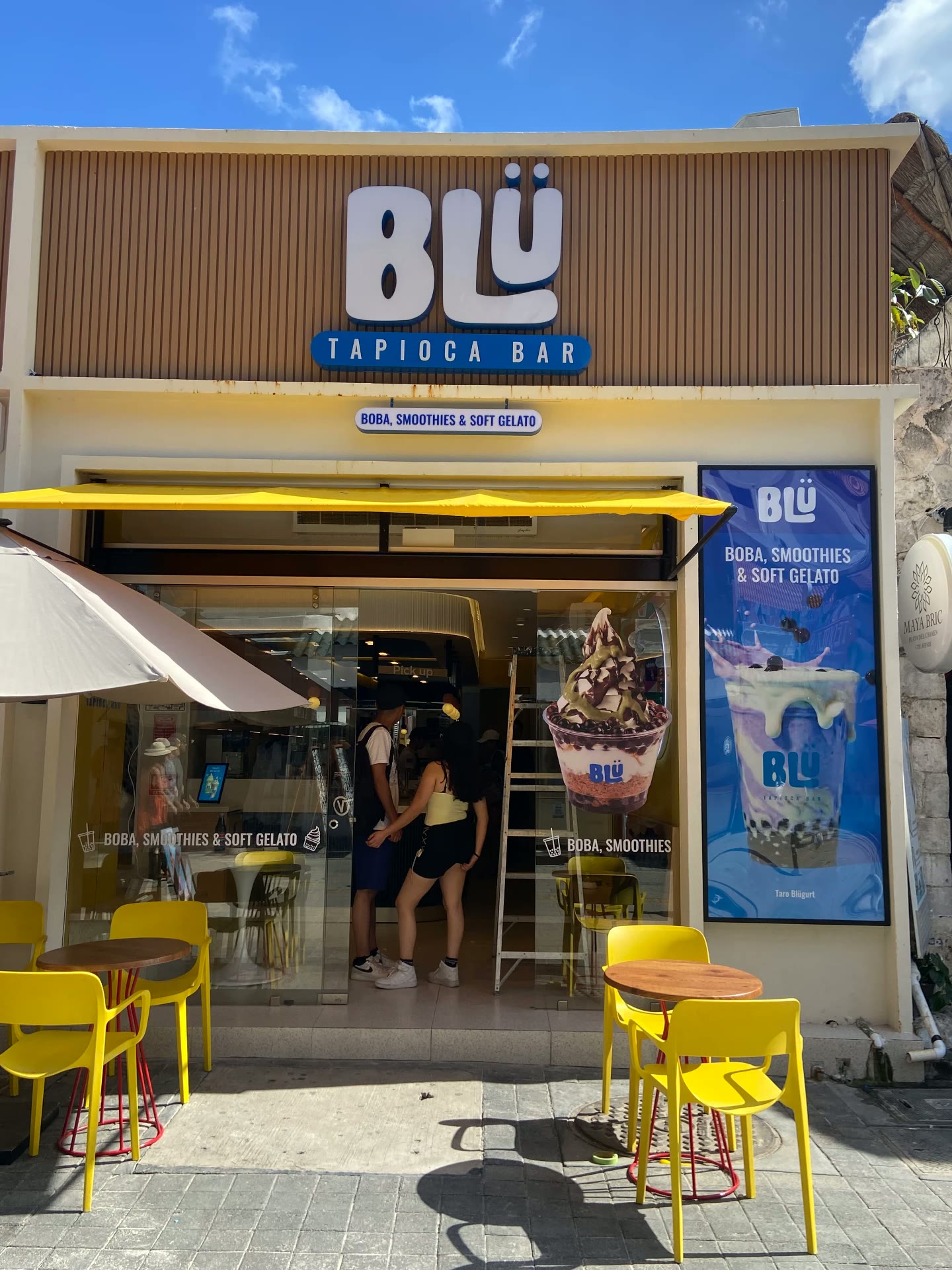 Fachada de BLÜ de día con terraza de sillas y mesas amarillas