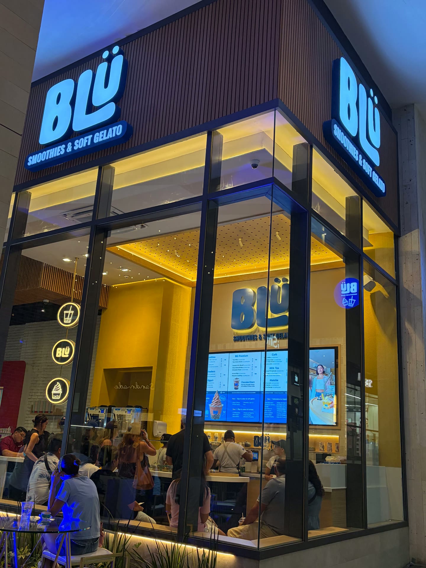 Fachada nocturna de BLÜ en esquina con letreros luminosos de Smoothies & Soft Gelato